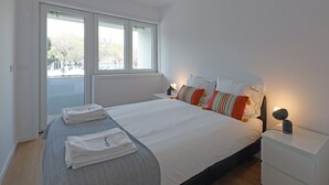 Ein ordentlich bezogenes Bett mit weißen Leinen, ein Nachttisch mit Lampe und ein Fenster mit Blick ins Freie.