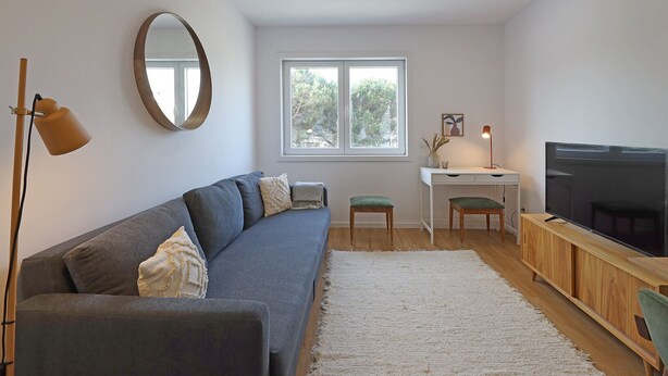 Ein modernes Wohnzimmer mit einem grauen Sofa, einem großen Spiegel, einem Fernseher und einem Schreibtisch mit Stuhl.
