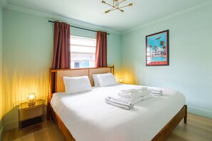 Una camera d'albergo con un letto grande, una finestra con tende, un quadro appeso al muro e un comodino con una lampada.