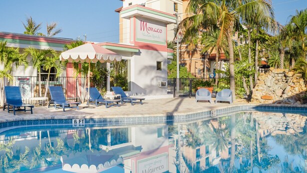 Un espace piscine avec des chaises longues, un panneau indiquant « Monaco Vacation Rentals », et des palmiers.