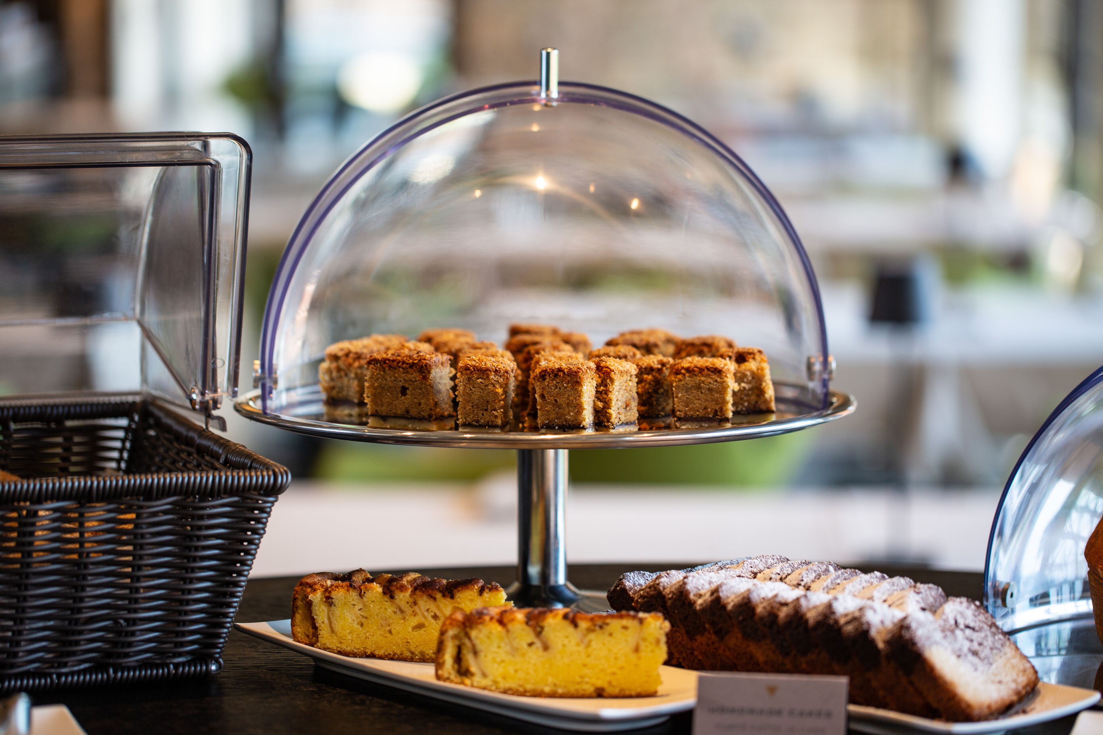 A display of various baked goods under a glass dome, including a cake and a loaf of bread, on a table with a wicker basket and a sign.