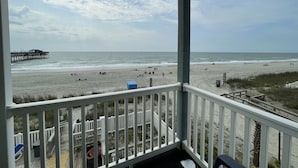 Udsigt over en strand fra en balkon med træterrasse, en mole i det fjerne og en sandstrand med mennesker og strandstole.