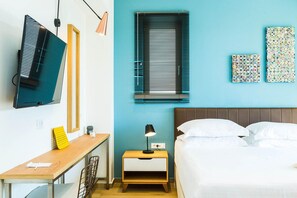A room with a bed, a desk, a TV, a nightstand with a lamp, and a window with blinds.