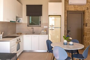 A compact kitchen with white cabinets, a built-in oven, a refrigerator, a window with a blind, and a dining area with a table and chairs. - Villa Vista I Free Heated Pool Dazzling Seaview (Kissamos)