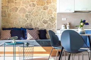 Un espace de vie avec un mur en pierre, un canapé agrémenté de coussins à motifs, une petite table ronde décorée d’un motif de poisson et une chaise bleue.