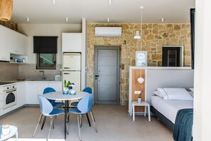 A compact kitchen with modern appliances, a dining area with blue chairs, a stone wall, a gray door, and a bed with white bedding. - Villa Vista I Free Heated Pool Dazzling Seaview (Kissamos)