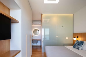 A modern hotel room with a bed, a TV, a desk, and a bathroom with a glass door.