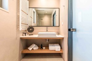 A modern bathroom with a large mirror, a white sink, and a wooden shelf with towels and toiletries.