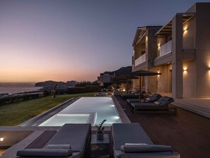 A modern poolside area with lounge chairs, a pool, and a view of the sea at dusk.