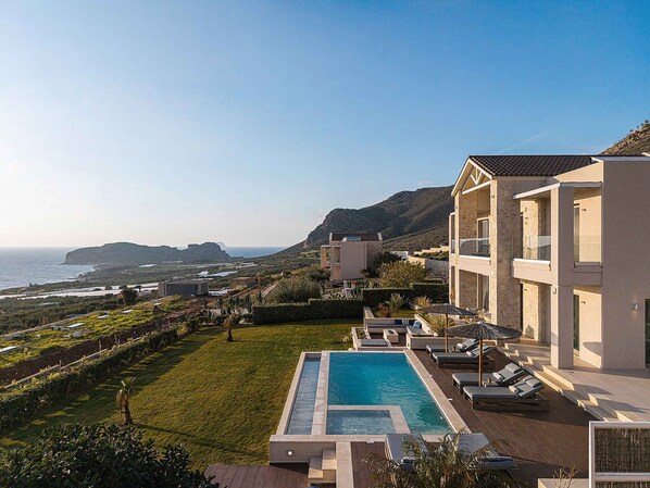A resort with a swimming pool, lounge chairs, and a view of the sea and hills.