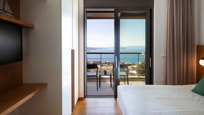 Une chambre d’hôtel avec un lit, un téléviseur posé sur une étagère en bois, un balcon avec une table et des chaises, et une vue sur la mer.