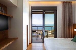 Une chambre d’hôtel avec un lit, un téléviseur posé sur une étagère en bois, un balcon avec une table et des chaises, et une vue sur la mer.