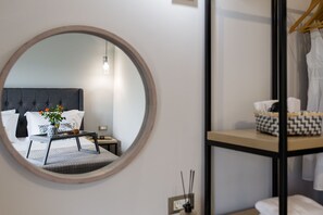 Un dormitorio con un espejo redondo, una cama con ropa de cama blanca, una mesita de noche con un florero y un perchero con ropa.