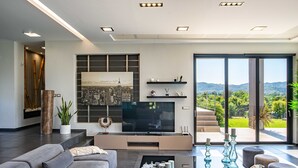 Un salon moderne doté d’un téléviseur à écran plat, de canapés gris, d’une table basse et d’une grande fenêtre donnant sur un paysage montagneux.