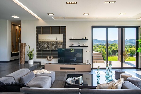 A modern living room with a flat-screen TV, grey sofas, a coffee table, and a large window offering a view of a mountainous landscape. - Villa Vega I Heated Pool Next to Nature (Platanias)