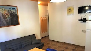 Ein Zimmer mit einer Couch, einem Tisch und Stühlen. An der Wand hängt ein Gemälde, und es gibt einen Fernseher sowie einen Kühlschrank.