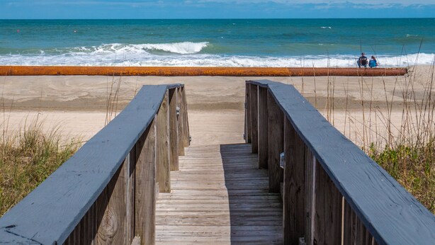 Een houten looppad dat naar een strand leidt, met een metalen hek en mensen die op het zand zitten.