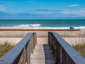 A wooden boardwalk leading to a beach with a metal barrier and people sitting on the sand.