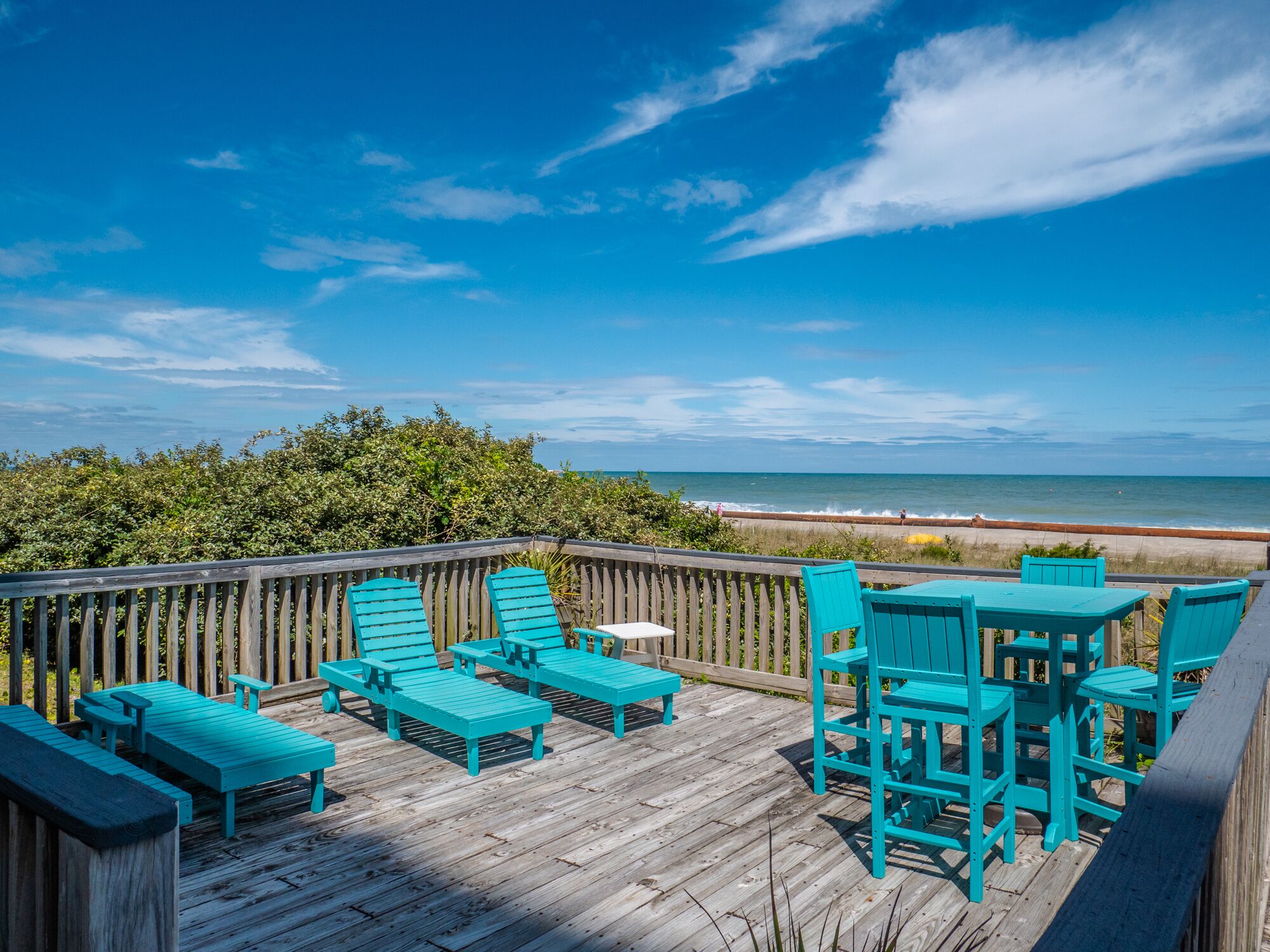 A wooden deck with turquoise outdoor furniture overlooking a beach.