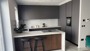 A modern kitchen with dark cabinets, a marble countertop, and a built-in stove. There are two bar stools and a potted plant on the counter. - Apartment 20 meters from the sea luxurious modern with all amenities (Tas-Sliema)