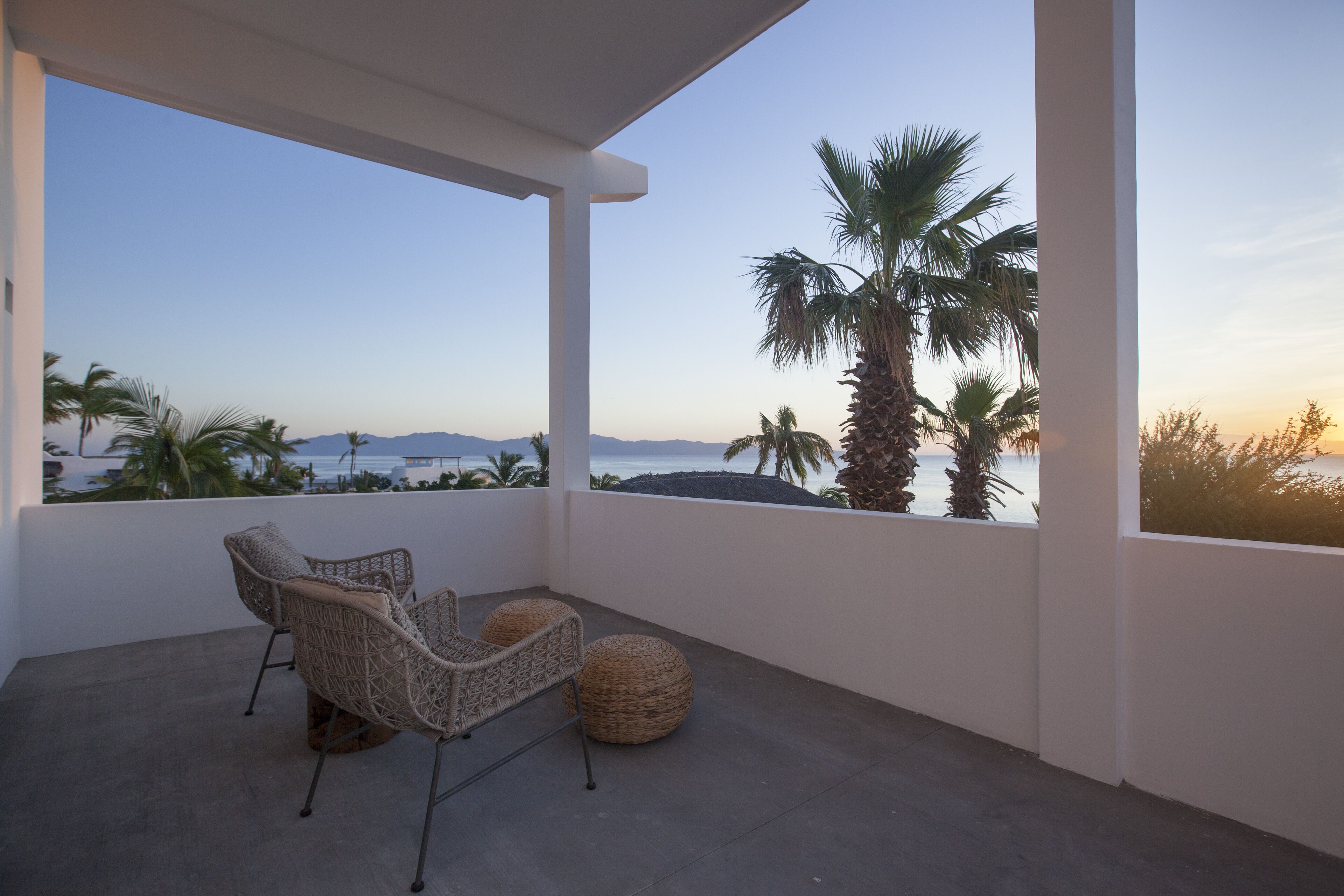 Un balcon agrémenté de fauteuils en osier et d’un pouf en fibres tressées, offrant une vue imprenable sur la côte, avec des palmiers et des montagnes en toile de fond.