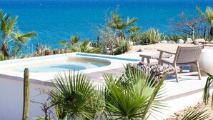 Zona de piscina con un jacuzzi, rodeada de plantas tropicales y con vistas a un mar azul cristalino y montañas lejanas.