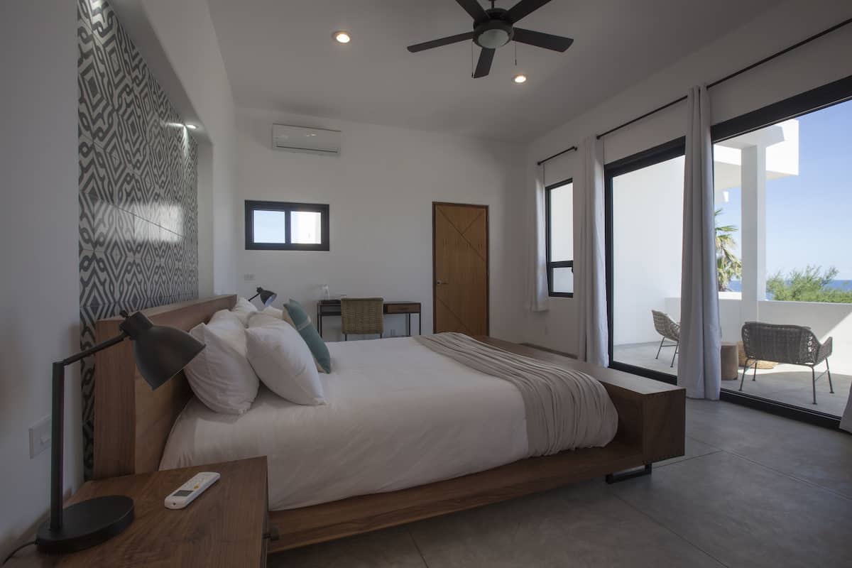 Ein modernes Schlafzimmer mit einem großen Bett, einem Deckenventilator, einem kleinen Tisch mit Lampe und Blick ins Freie durch große Fenster.