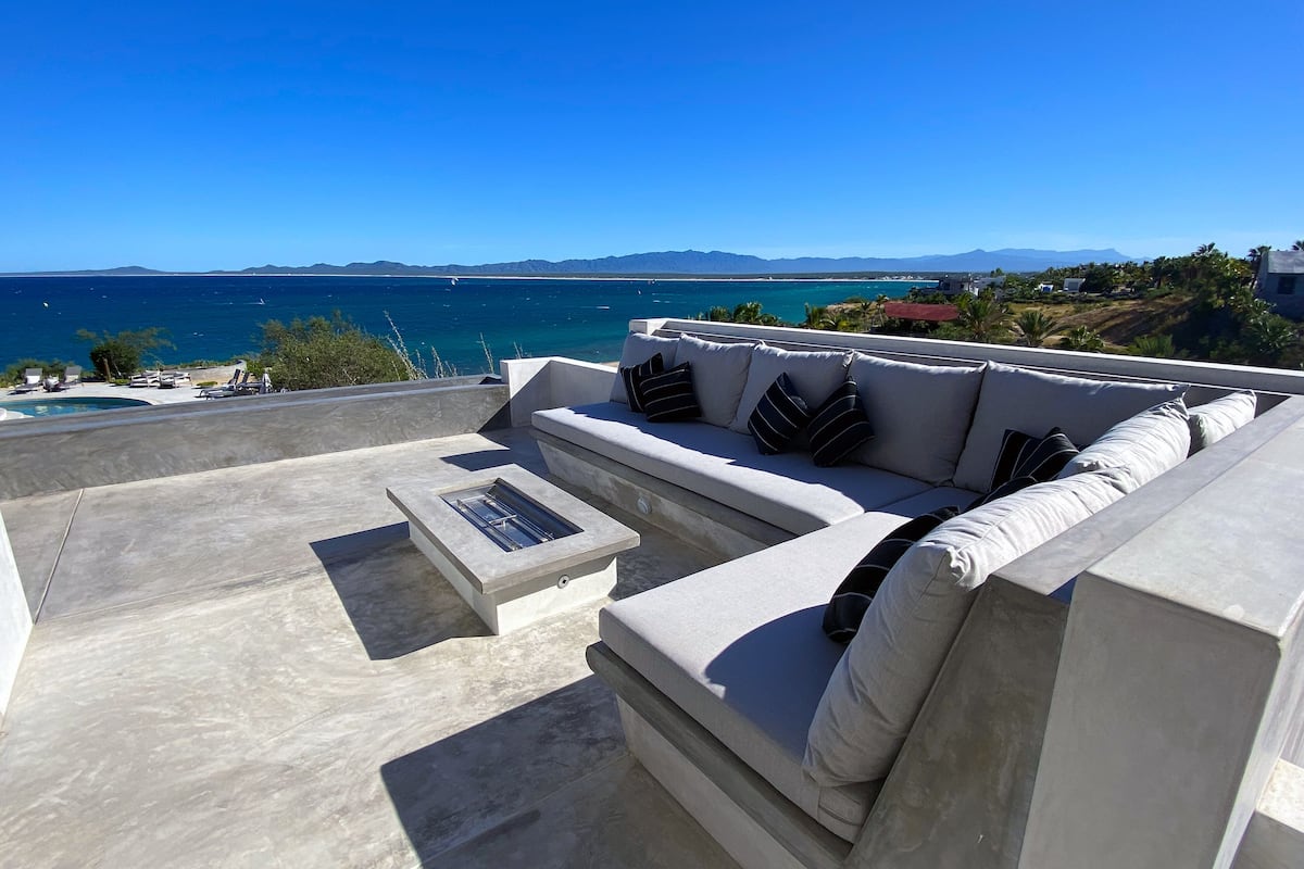 Ein moderner Außenbereich mit einer Sitzecke, einer Couch und einem Tisch, mit Blick auf die Küste und Berge in der Ferne.