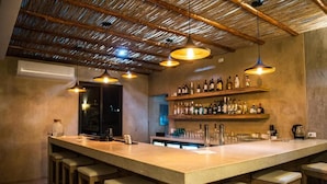 Una zona de bar con una barra larga, taburetes, lámparas colgantes y estantes con botellas.
