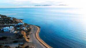 Vista aérea de una zona costera con playa, un pequeño poblado de edificios blancos y una vista despejada del mar y montañas distantes.