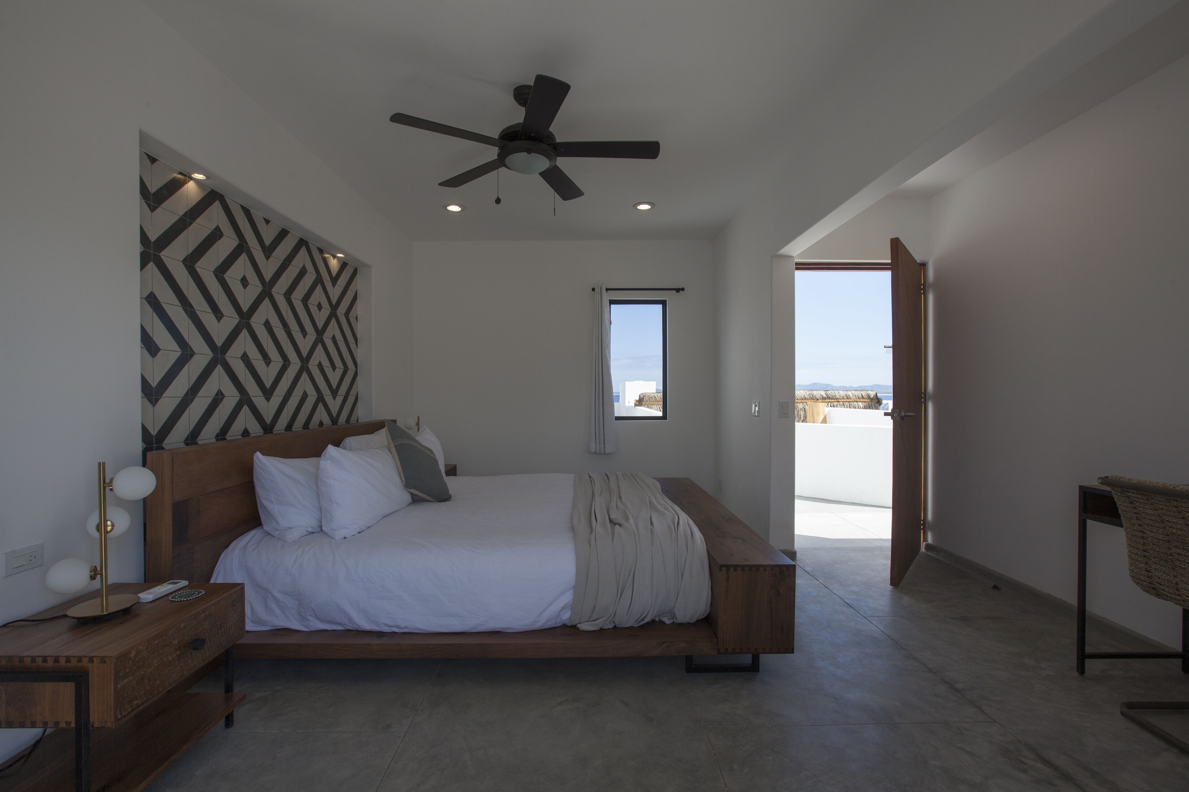 Quarto moderno com uma cama grande, um ventilador de teto, uma mesa de cabeceira com abajur e vista para o exterior pela janela.