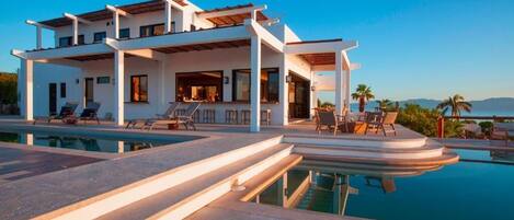 Una casa moderna de dos pisos con piscina, mobiliario exterior y vista al mar.
