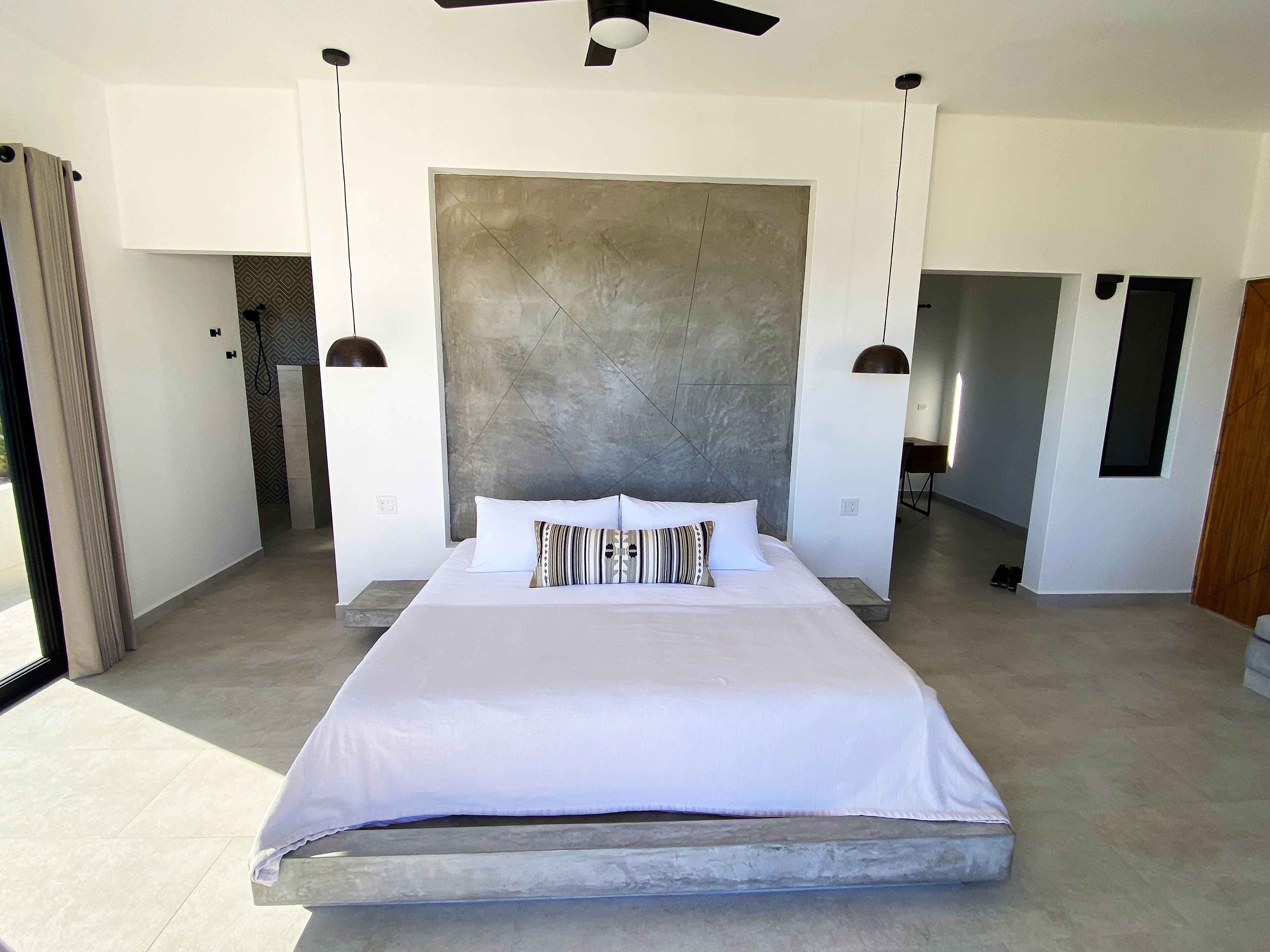 Un dormitorio moderno con una cama grande, una pared de concreto como detalle decorativo e iluminación colgante.