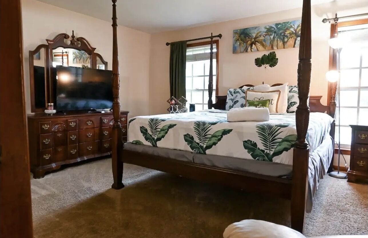 A four-poster bed with a leaf-patterned bedspread, a wooden dresser with drawers, a flat-screen TV, and a painting of palm trees on the wall.