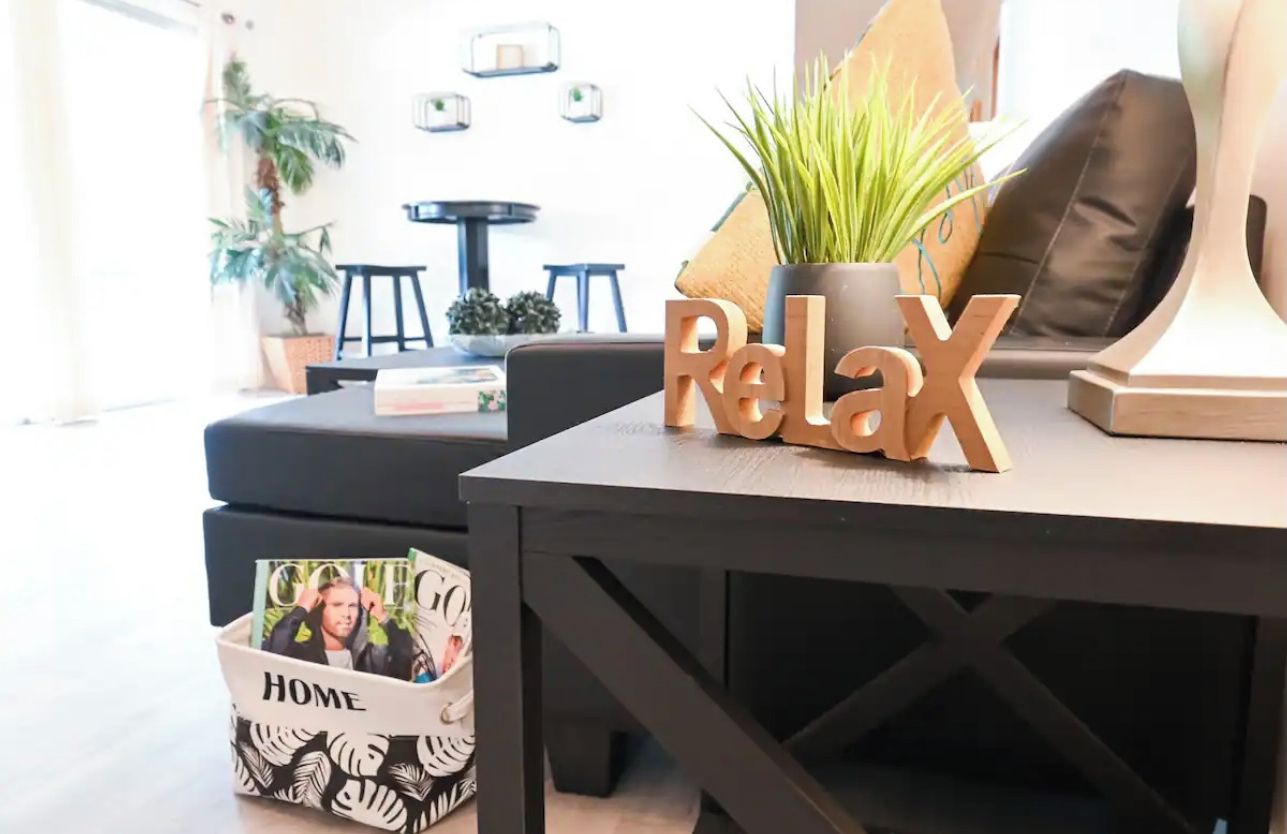 A modern living room with a dark wooden table, a potted plant, and a 'Relax' sign. There is a small black and white patterned basket labeled 'HOME' on the floor.
