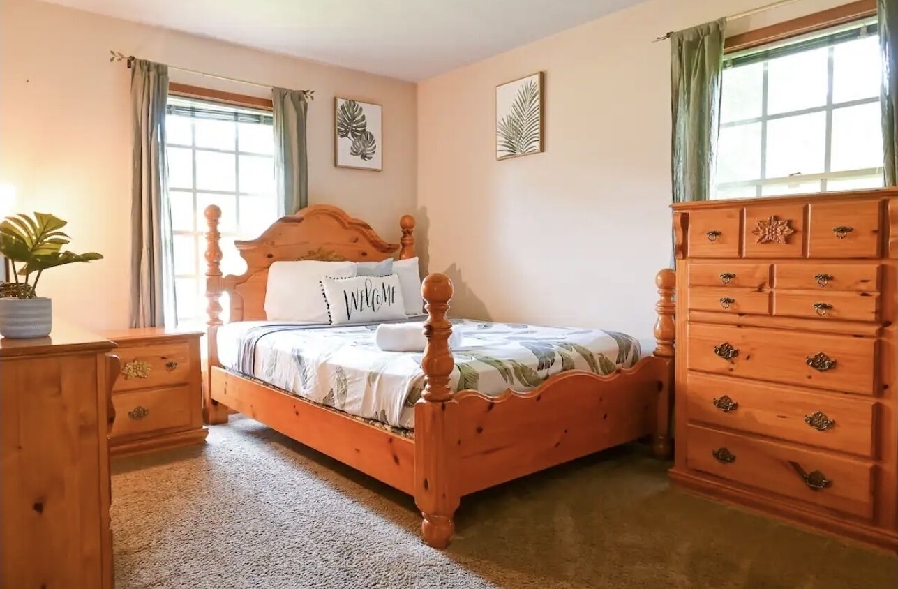 Quarto com cama de madeira, mesa de cabeceira, penteadeira e janela com cortinas.