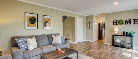 A living room with a gray sofa, a coffee table, and a rug. There are framed pictures on the wall and a 'HOME' sign.