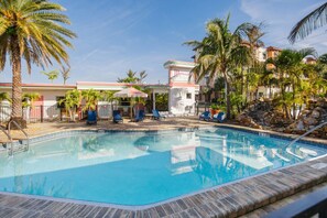 A swimming pool with lounge chairs and umbrellas, surrounded by palm trees and a building with a sign.