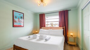 Een netjes opgemaakt bed met witte lakens en handdoeken, een ingelijst schilderij aan de muur, een raam met rode gordijnen en een klein nachtkastje met een lamp.