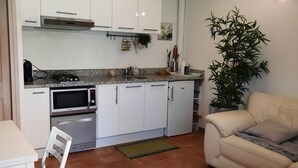 Cozinha compacta com armários brancos, micro-ondas e fogão. Há uma mesa de jantar com cadeiras e um sofá na área de estar. Uma planta em vaso é visível no canto.