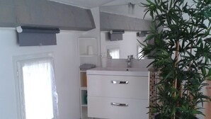 Casa de banho com toucador branco, lavatório, espelho, planta em vaso e um pequeno contentor de lixo.
