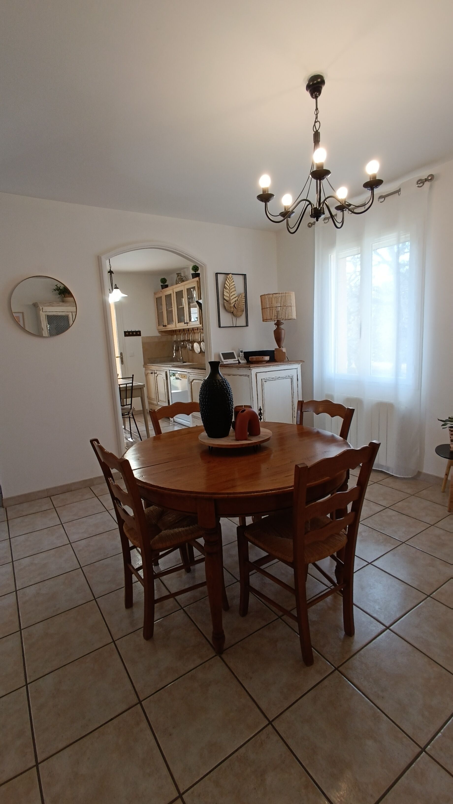 A dining area with a round wooden table, six chairs, a chandelier, and a kitchen in the background.