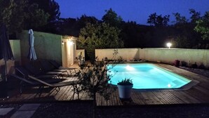 A rectangular hot tub surrounded by a wooden deck, with lounge chairs and a small covered area with lights.