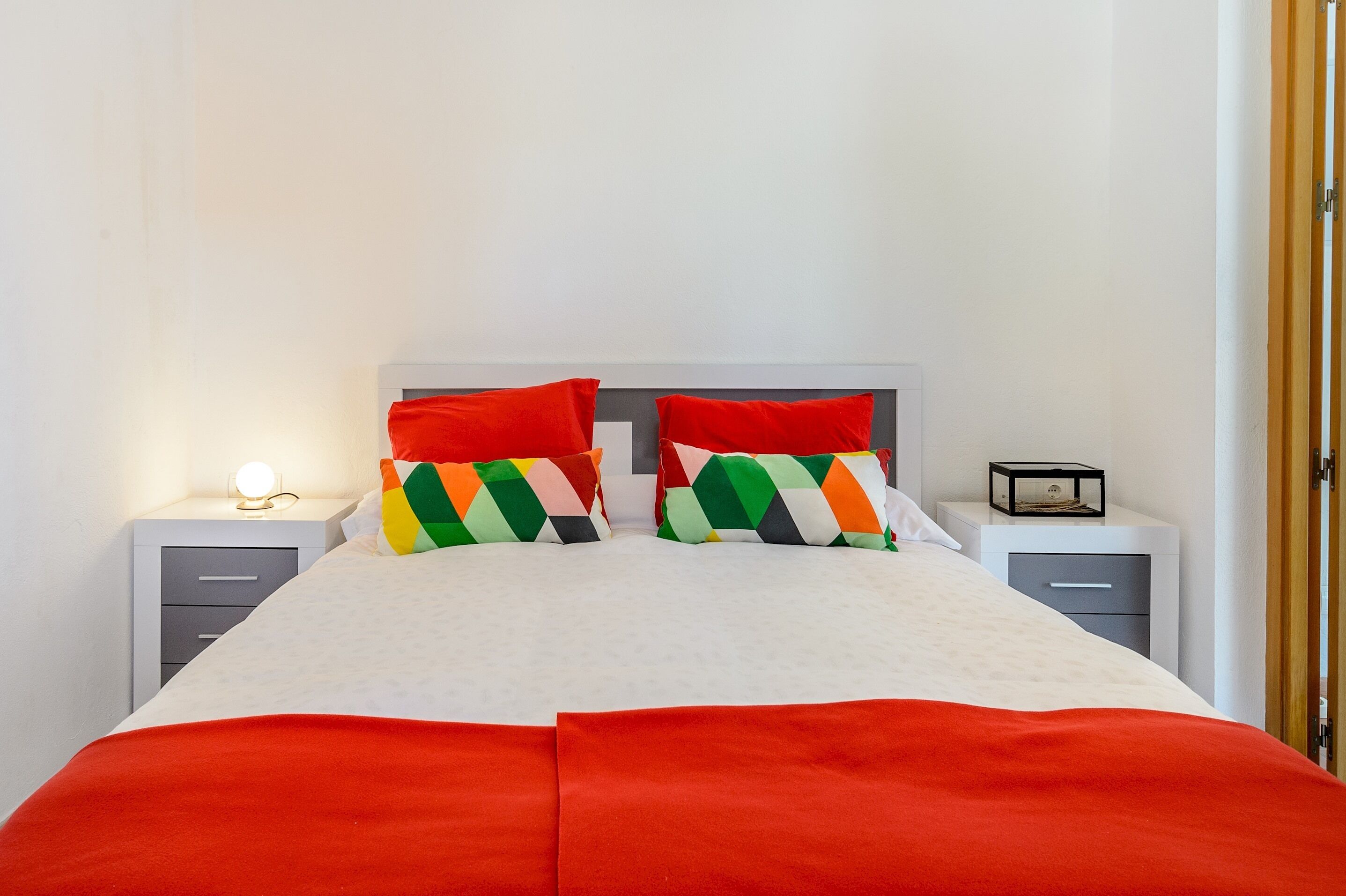 Ein ordentlich bezogenes Bett mit einer weiß-roten Tagesdecke, zwei farbenfrohen Kissen und einer Nachttischlampe. Es gibt zwei Nachttische, einer mit einer Lampe und der andere mit einer kleinen Uhr.