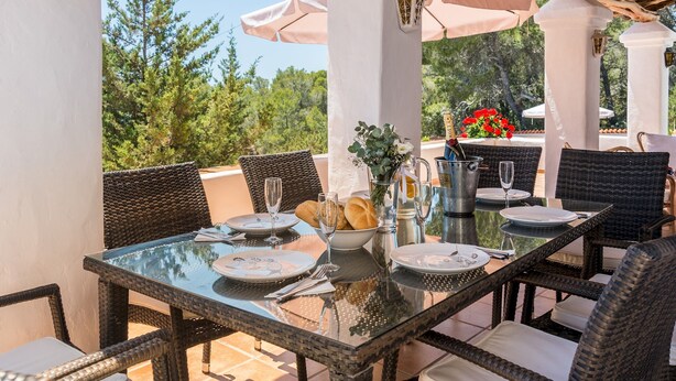 An outdoor dining area with wicker chairs and a glass table, surrounded by white pillars and umbrellas, set against a backdrop of trees and a clear sky. - Villa Las Tres Yucas (Sant Antoni de Portmany)