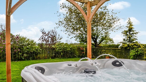 Een hottub met een houten pergola erboven, omgeven door een weelderige tuin en een houten omheining.