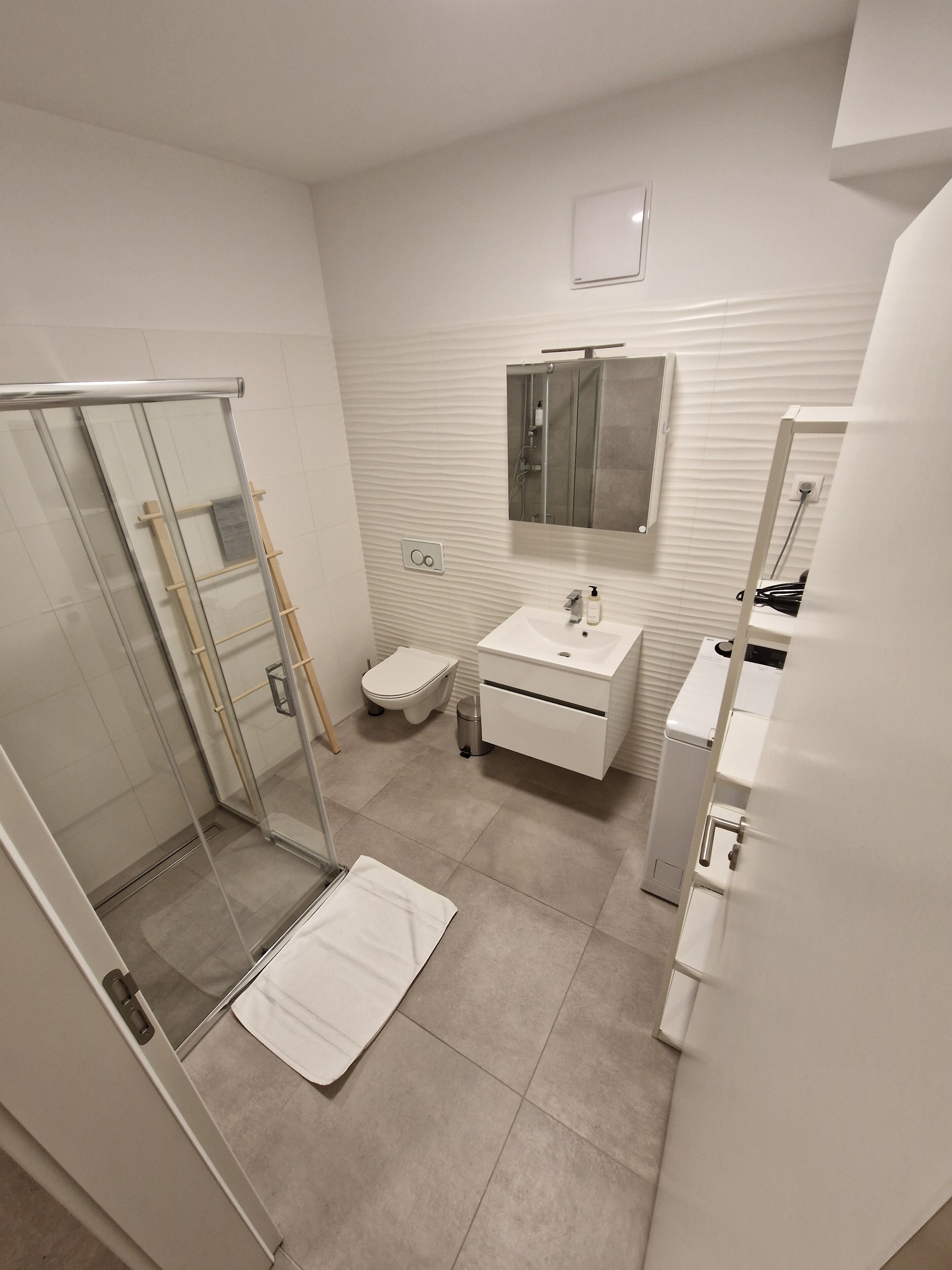 Ein modernes Badezimmer mit Duschkabine aus Glas, Toilette, Waschbecken mit Spiegel und Handtuch auf dem Boden.