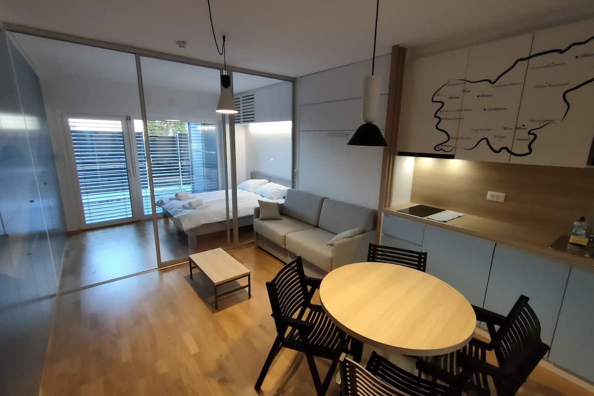 A modern, well-lit room with a wooden floor, a round dining table with chairs, a sofa, a bed with white bedding, and a map on the wall.