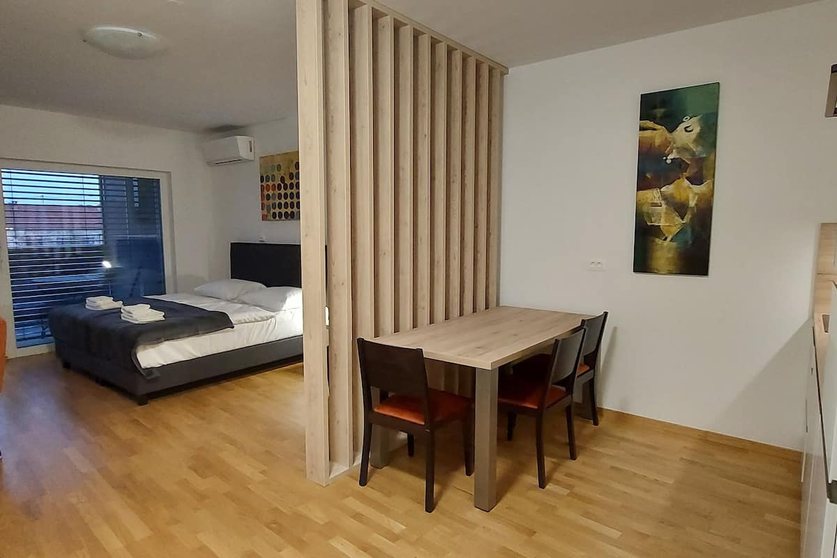 A bedroom with a bed, a desk, and a chair. There is a window with blinds, a painting on the wall, and a wooden floor.