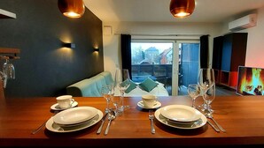 A dining area with a wooden table set for four, including plates, cutlery, and wine glasses. There is a sofa with cushions in the background, and a television displaying a fire scene is mounted on the wall. - REZIDENCA PIPANOVA (Ljubljana)